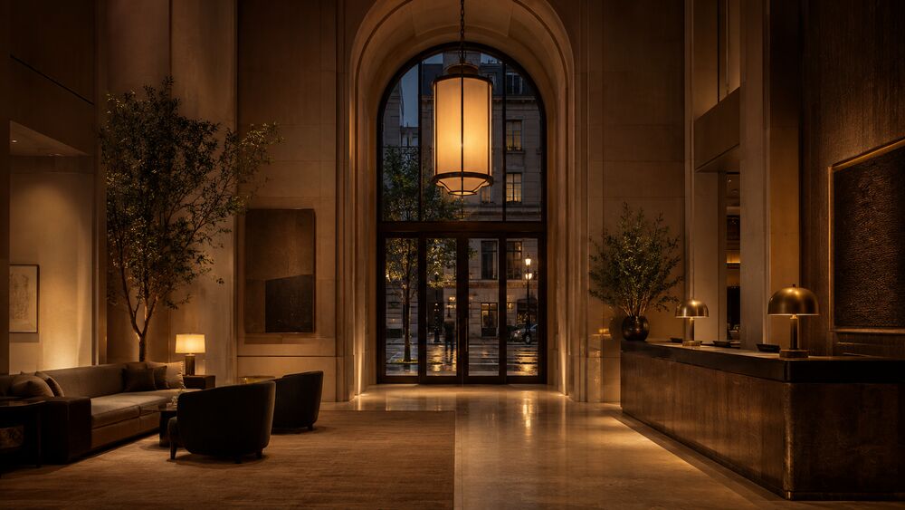 The interior lobby seen from just inside the arched entry doors at dusk — honed-limestone walls, a single oversized warm pendant lantern, a low front desk in dark bronze and stone, and an ikebana arrangement on a stone console.